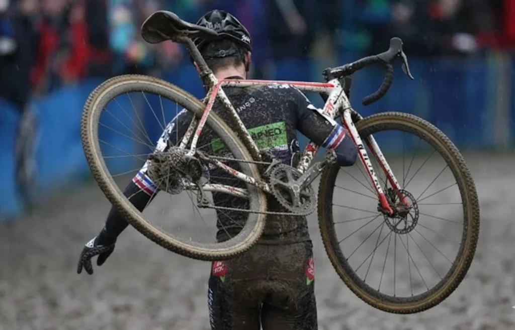 Ciclista en competencia de ciclocross cargando su bicicleta sobre el hombro en un circuito lleno de barro